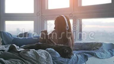 年轻可爱的女士戴着耳机，和她可爱的缅因州猫在床上放松，听着窗户边的音乐唱歌